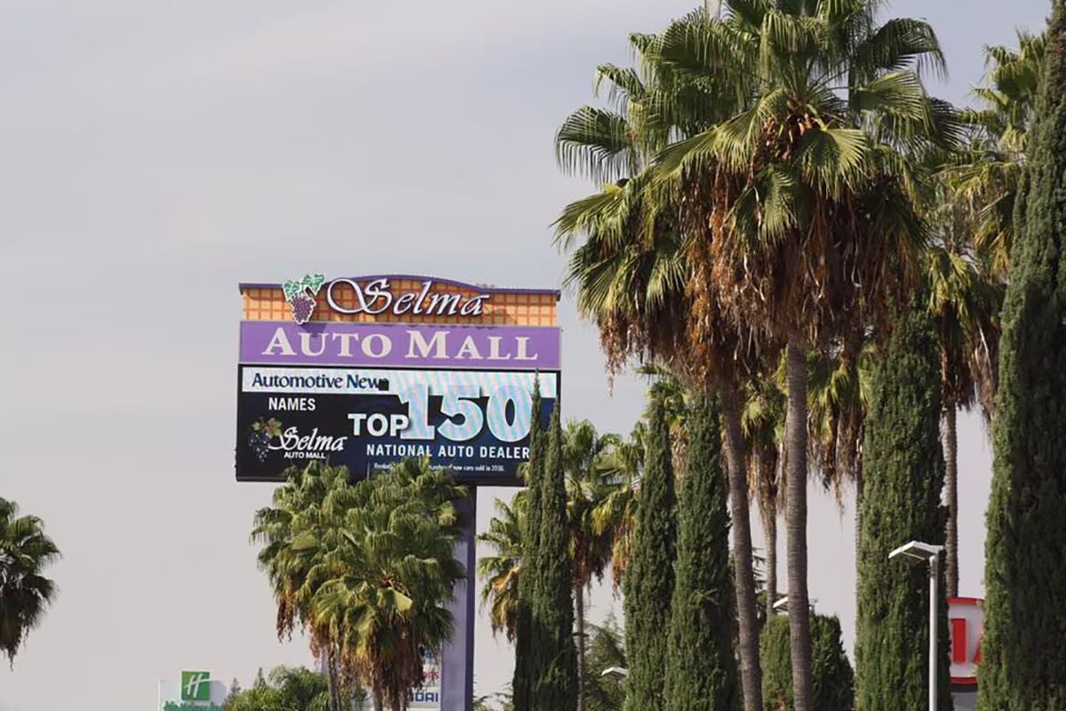 'Selma Auto Mall' sign with 'Top 150 National Auto Dealer' against a sky, surrounded by palm and cypress trees.