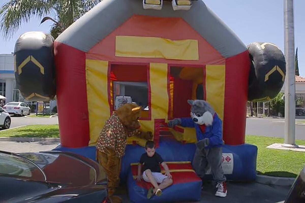 Two mascots, a bear and a wolf, interact with a young boy sitting on a red and blue inflatable in front of a bouncy house.