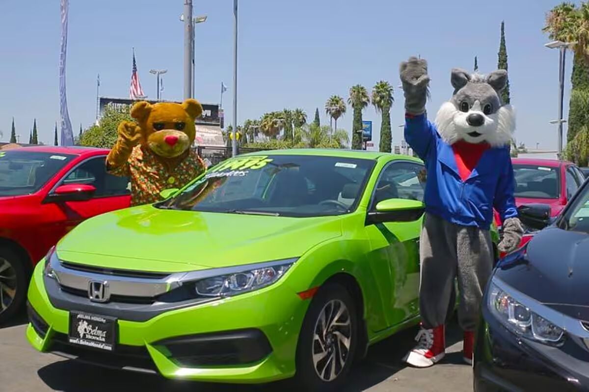 Two people in mascot costumes, a brown bear and a gray wolf, stand beside a lime-green Honda car.